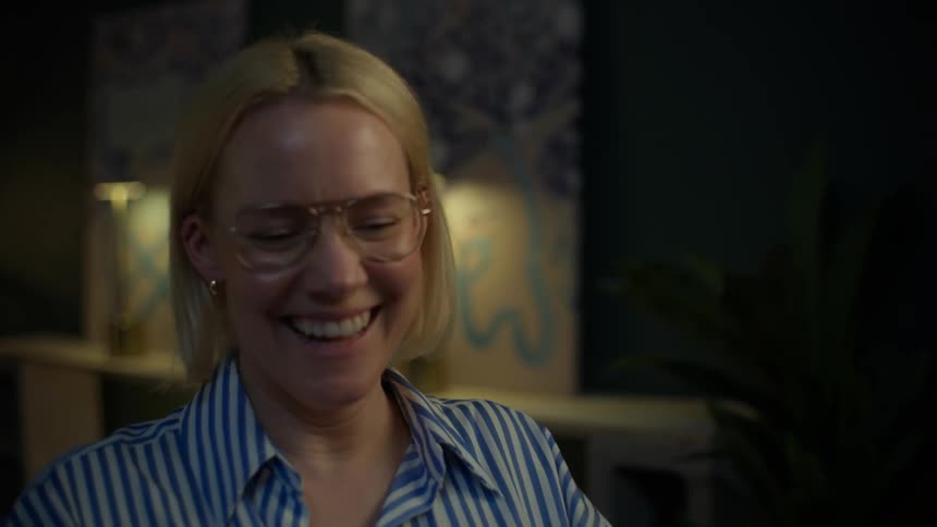A slow-motion close-up of a joyful blonde woman with glasses bursting into laughter in a cozy room