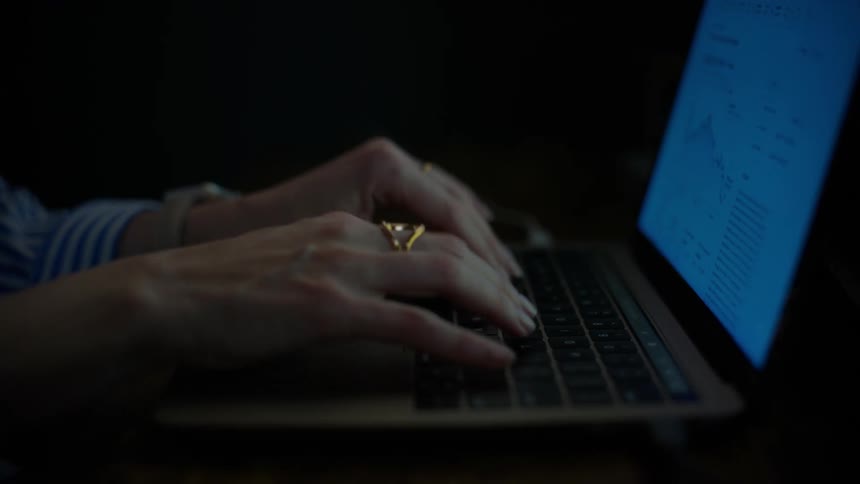 Slow motion close-up of a woman typing on her laptop keyboard in a dark room, with the screen light illuminating her hands