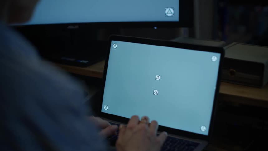 An over-the-shoulder shot of a person working at a desk on a laptop with a trackable screen, tilting down from an external monitor