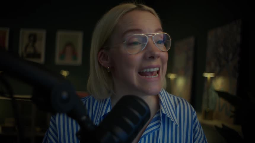 An expressive young woman with glasses records a podcast or live stream, speaking passionately into a professional microphone in her stylish, dimly lit home studio