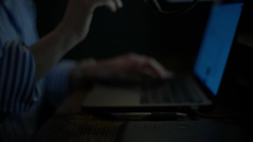 A woman types on a laptop and then speaks into a microphone while recording a podcast in a dark studio