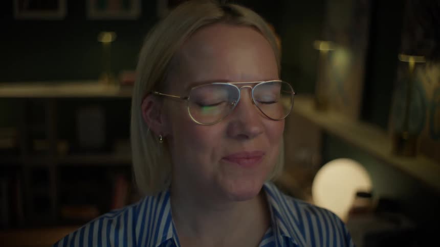 A woman with glasses smiles and laughs while working on her computer late at night in a dimly lit home office