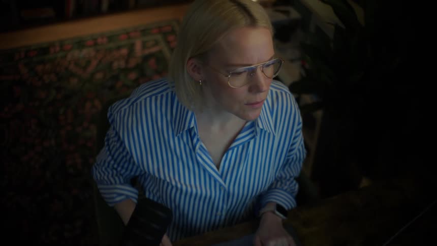 A professional woman wearing glasses works late on her laptop in her home office, her expression shifting from concentration to a subtle smile