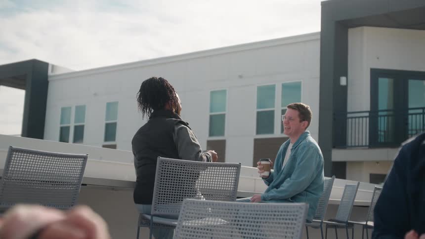 Friends socialize and laugh while enjoying coffee on a sunny modern rooftop patio in an over-the-shoulder view