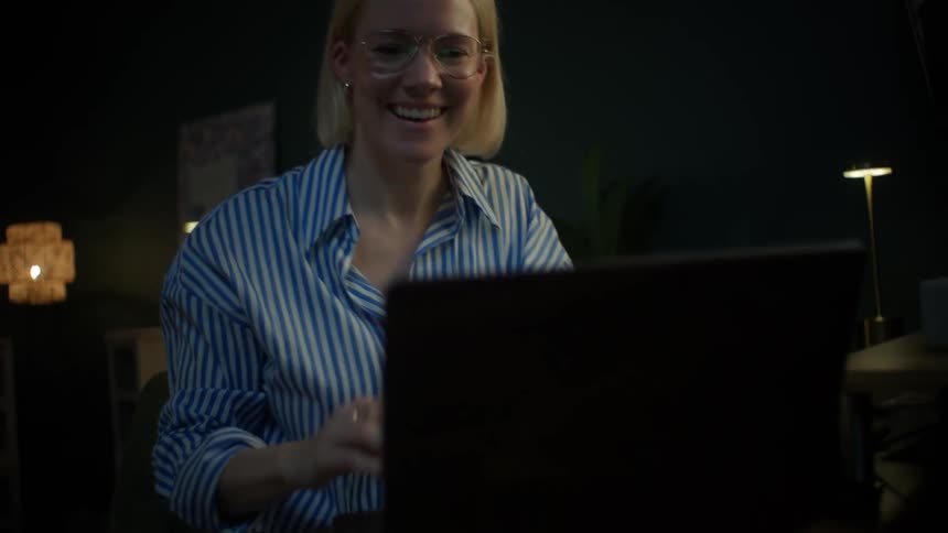 A smiling woman with glasses opens her laptop at her desk in a cozy, dimly lit room at night