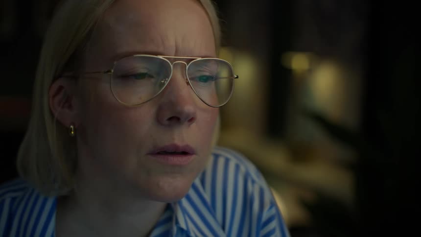 A worried blonde woman in glasses stares intently at a screen, her face illuminated by its glow in a dark room