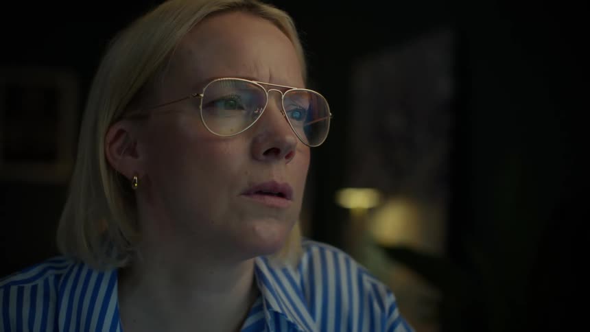 A focused blonde woman wearing glasses works late into the night, her serious expression illuminated by the glow of her computer screen