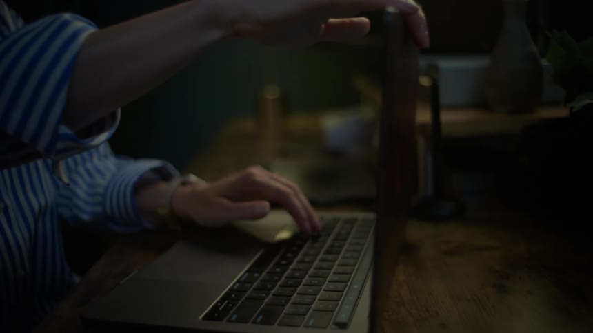 A woman types on her laptop at a wooden desk in a dimly lit room and then slowly closes the lid when she is finished