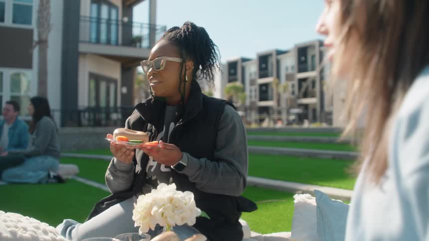Two young women chat and laugh while enjoying a picnic on a beautiful sunny day in a modern apartment courtyard