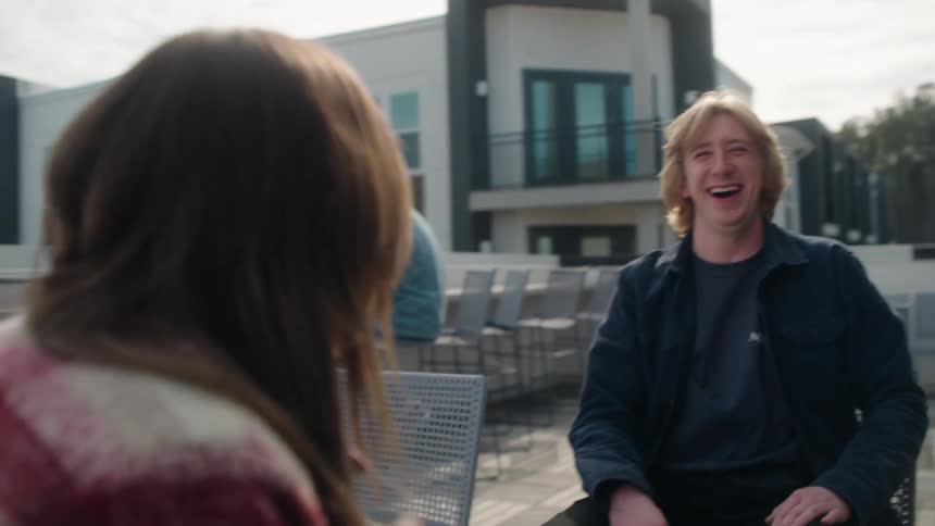 A slow-motion over-the-shoulder shot reveals a young man and woman laughing and talking together on an apartment rooftop patio