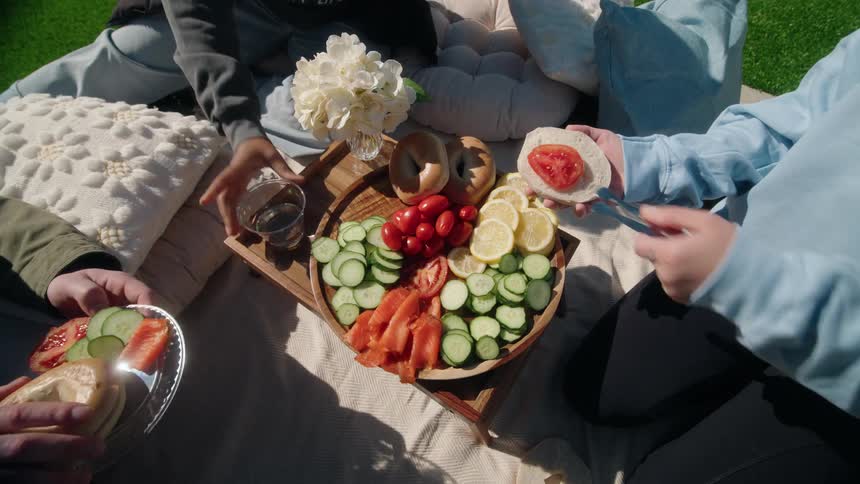 A group of friends enjoys a sunny outdoor picnic, preparing bagels with a colorful platter of lox and fresh vegetables