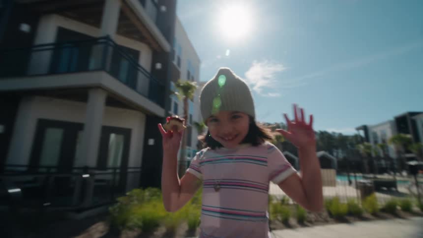 A happy young girl in a beanie roller skates in slow motion through a modern apartment community on a sunny day, smiling at the camera