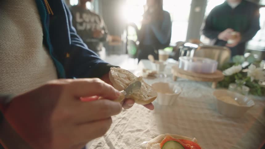 A person spreads cream cheese on a fresh bagel at a sunny brunch table surrounded by friends