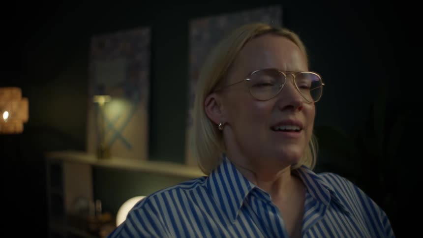 A smiling blonde woman wearing glasses closes her laptop, finishing her work for the night in a warmly lit room