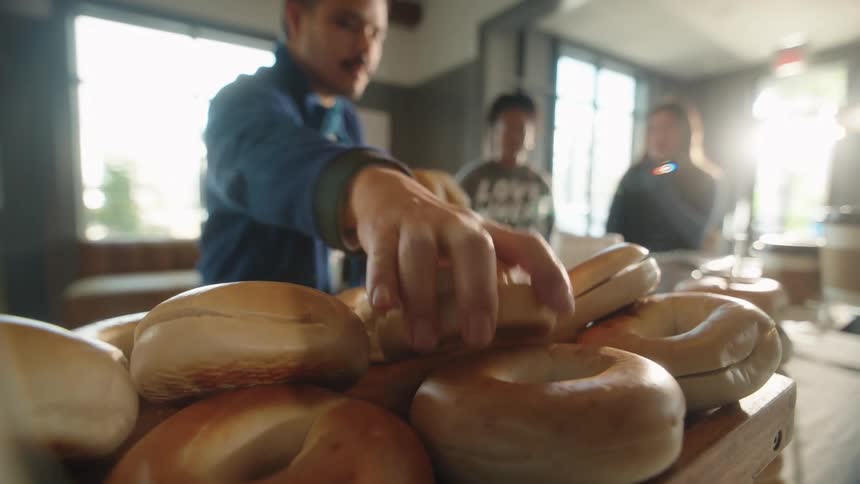 A close-up slow-motion shot of fresh bagels on a wooden board as a man selects one in a sunlit cafe