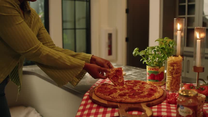 A woman takes a cheesy slice of pepperoni pizza from a candlelit Italian food spread in a cozy kitchen
