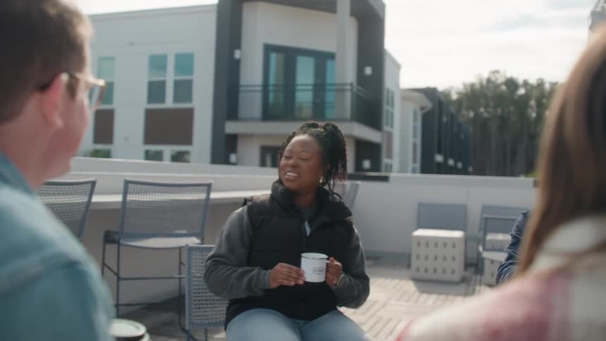 A diverse group of young adult friends socialize over coffee on an apartment rooftop patio on a sunny day