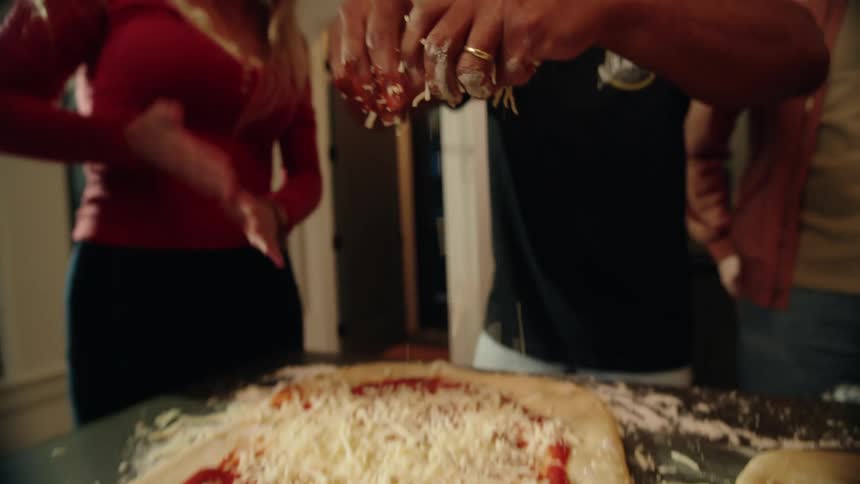 A diverse group of friends laugh and have fun while sprinkling shredded cheese onto a homemade pizza in a kitchen