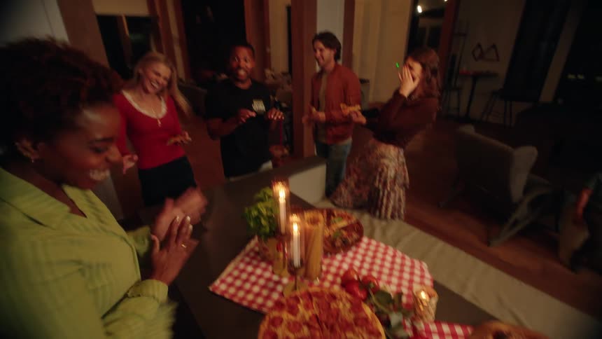 A diverse group of friends laugh together at a pizza party, with the camera tilting down to the food in slow motion