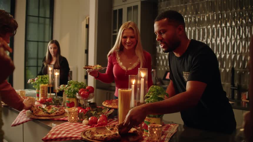 A diverse group of friends laugh and serve themselves fresh pizza at a cozy, candlelit dinner party in a modern kitchen