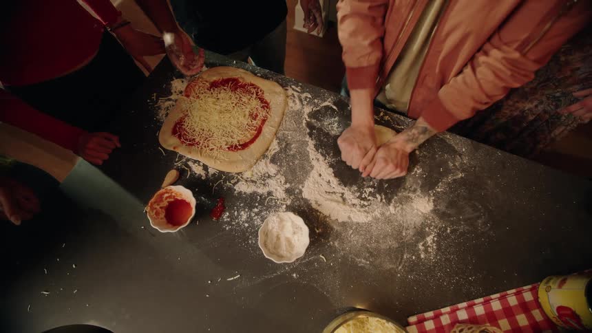 From above, a group of friends gathers in a kitchen to make homemade heart-shaped pizzas together