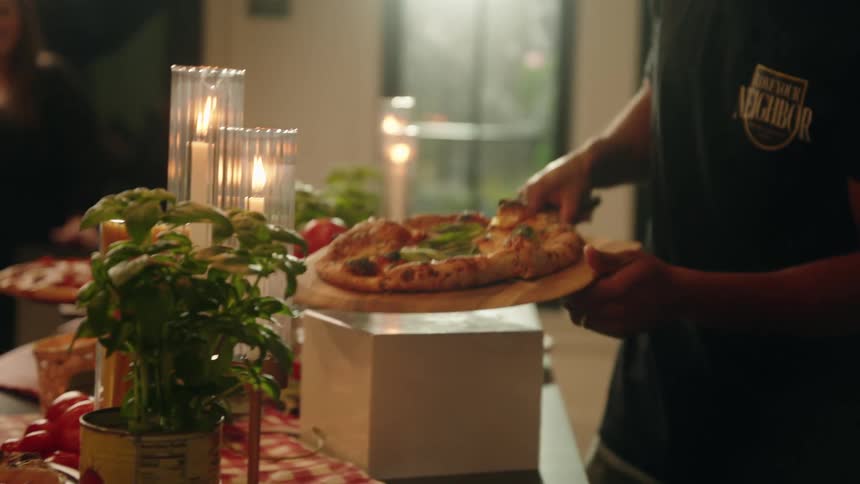 A man carefully brings a hot, freshly baked pizza on a wooden peel to a candlelit dinner table in slow motion