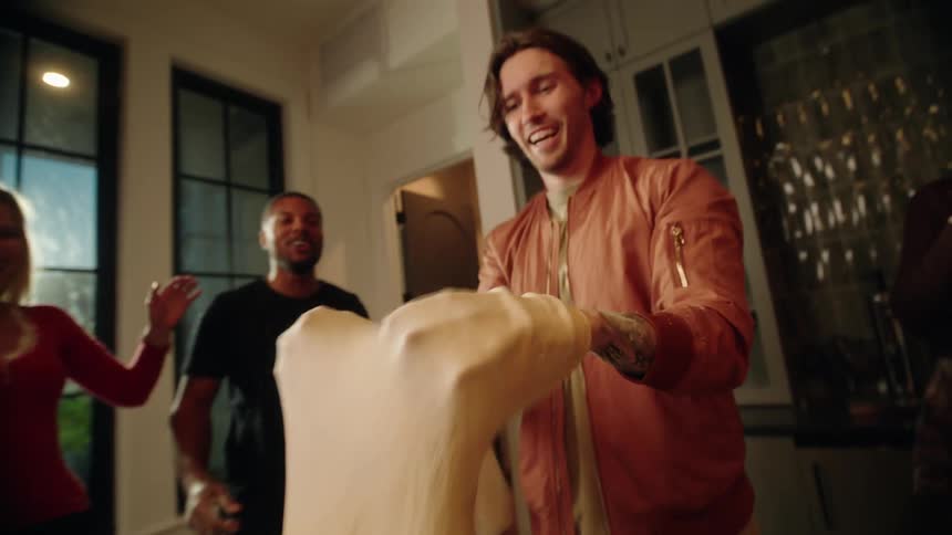 A young man tosses pizza dough in the air in slow motion while his impressed friends watch and laugh during a party in the kitchen