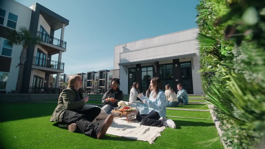 A diverse group of young friends enjoys a sunny picnic, talking and toasting on the green lawn of a modern apartment courtyard