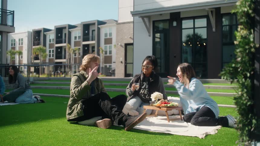 A group of young, diverse friends share a laugh and a charcuterie board during a picnic on the lawn of a modern apartment building