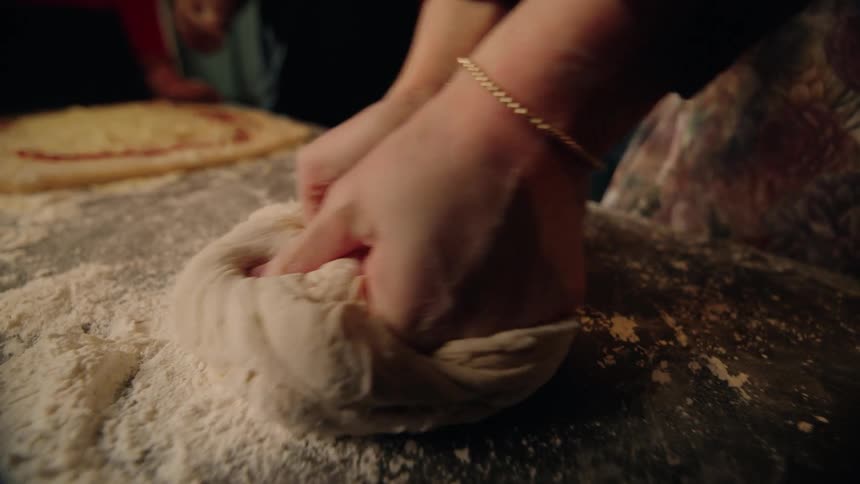 Close-up slow motion shot of a woman's hands kneading pizza dough on a floured surface while friends watch
