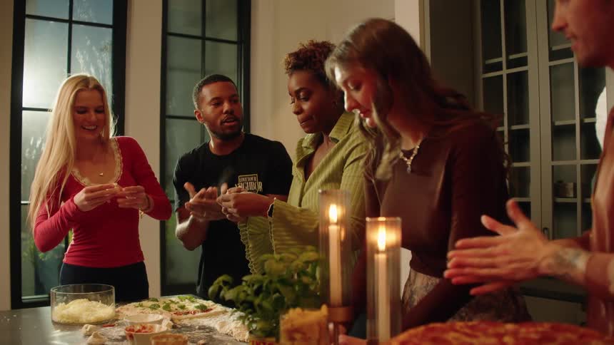 Happy and diverse friends laugh and talk while making homemade pizza together during a fun dinner party in a cozy kitchen