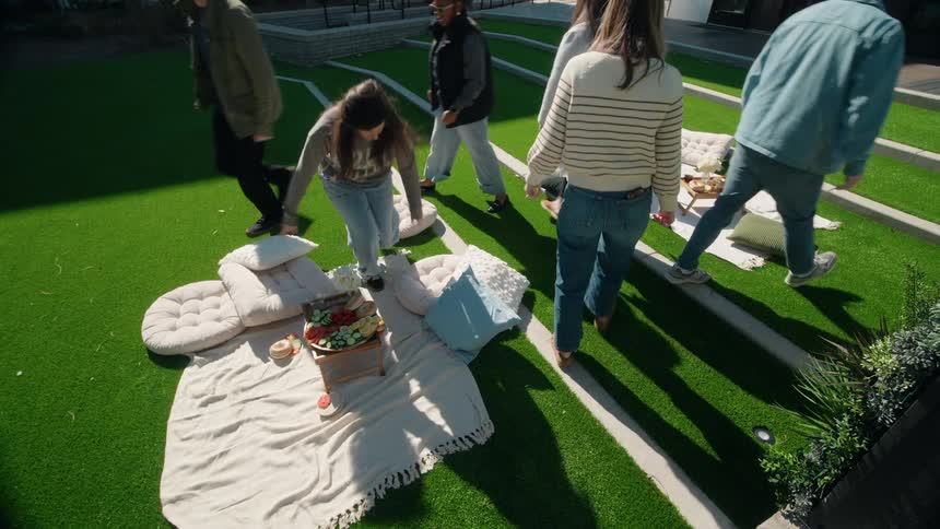 Two groups of diverse friends enjoy a sunny day picnic, talking and sharing food on a modern terraced grass lawn
