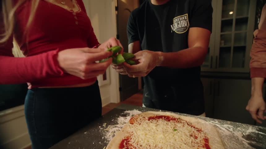 A group of diverse friends gather in a kitchen to make homemade pizza together, adding fresh basil as a topping