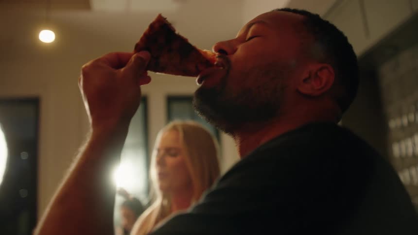A man enthusiastically eats a slice of pizza in slow motion, closing his eyes to savor the delicious taste