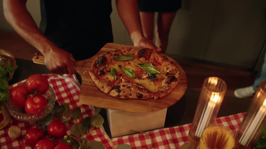 A man carefully places a freshly baked homemade pizza on a wooden peel onto a beautifully decorated dinner table in slow motion