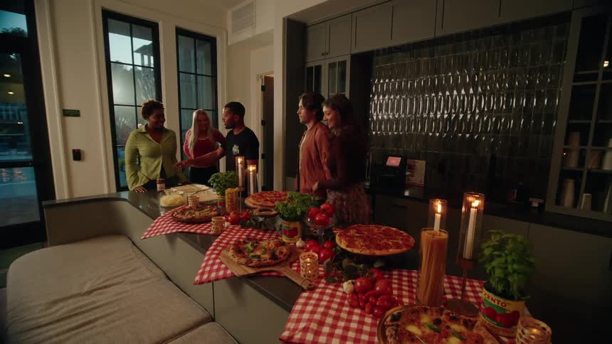 An excited host welcomes his diverse group of friends to a festive, candlelit pizza party in a modern kitchen