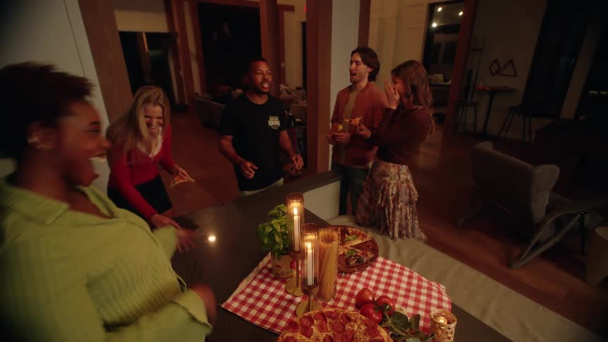A group of diverse friends laugh and socialize at a cozy, candlelit pizza party in a modern home