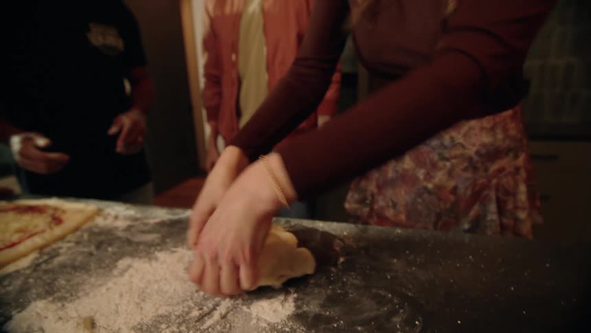 A woman kneads homemade pizza dough on a floured countertop while her friends watch and smile during a gathering
