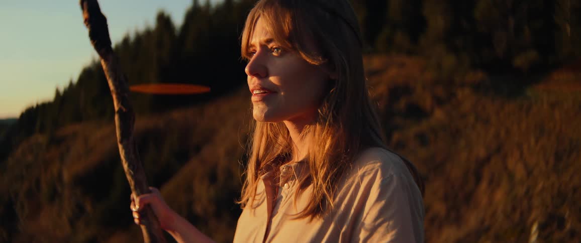 A serene young woman holding a wooden staff enjoys the beautiful golden hour light while looking out over a forested mountain landscape