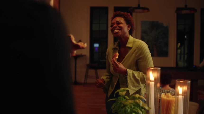 A diverse group of friends and family eat pizza, laugh, and socialize during a cozy candlelit dinner party at home