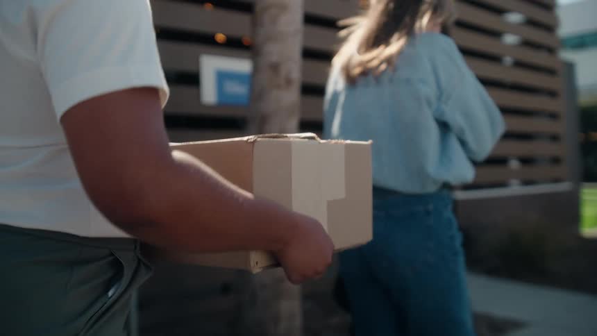 A person carries a cardboard box while following a woman down a sidewalk in slow motion on a sunny day