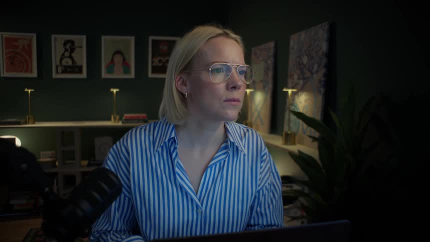 A focused woman wearing glasses looks intently at her computer screen while working late in her dark, stylish home office