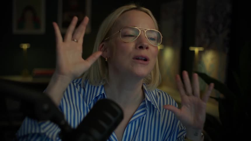 A blonde woman with glasses passionately records a podcast, speaking animatedly into a microphone in a dimly lit home studio in slow motion