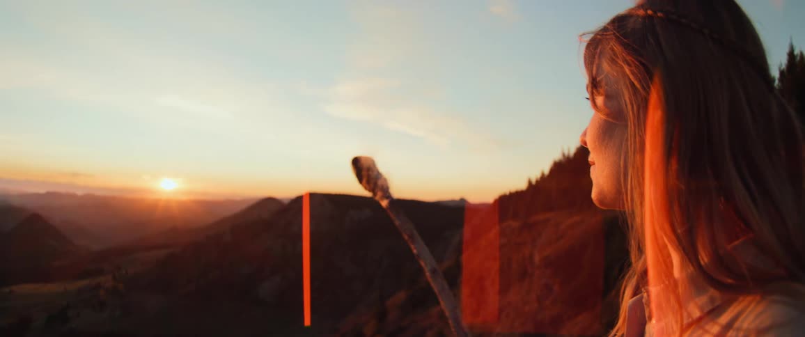 A serene woman with a walking staff watches the beautiful golden sunset over a vast mountain valley