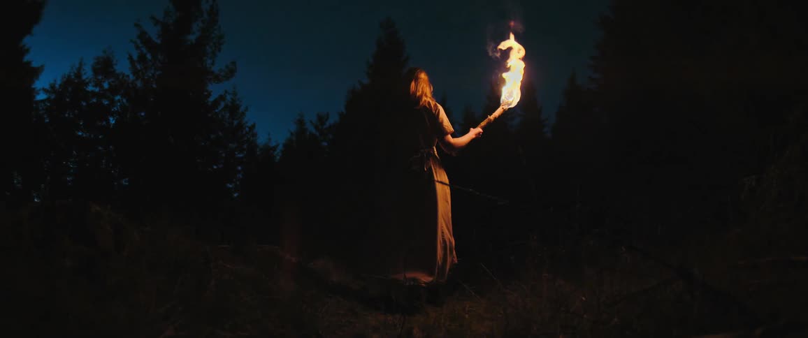 A woman in historical attire walks into a dark forest at night holding a flaming torch