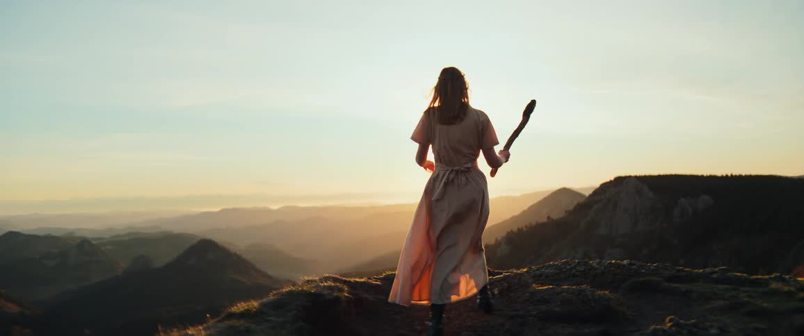 A woman in a flowing dress walks toward the sunrise on a mountain peak, holding a staff in this cinematic slow-motion shot