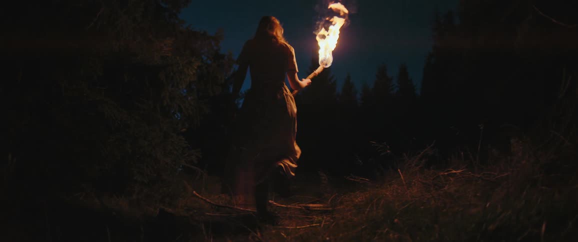 A woman holding a flaming torch walks into a dark forest at night in a dramatic slow-motion shot