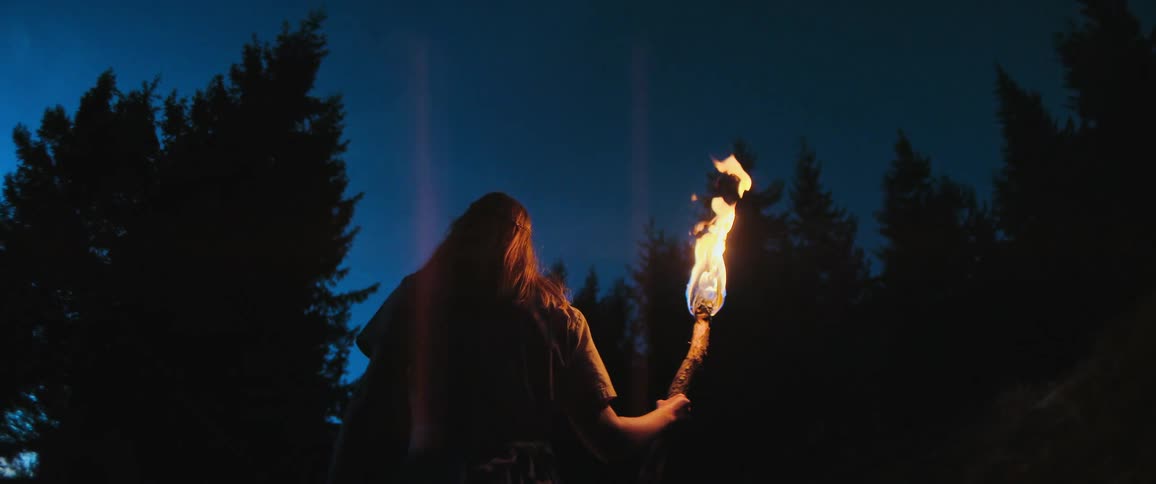 A woman with long hair stands silhouetted against a dark blue sky, holding a burning torch in a forest at night