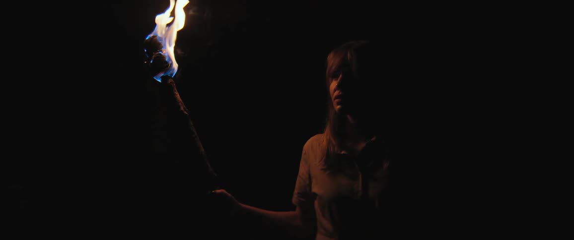 A tense woman holds a burning torch, her face illuminated by the flame as she stands in the pitch-black darkness