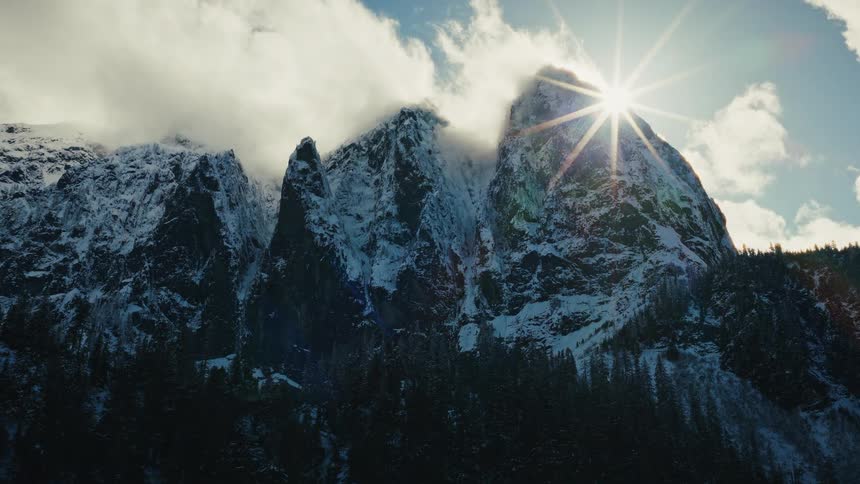 The sun rises from behind a majestic, snow-covered mountain peak, creating a brilliant starburst in the cloudy sky